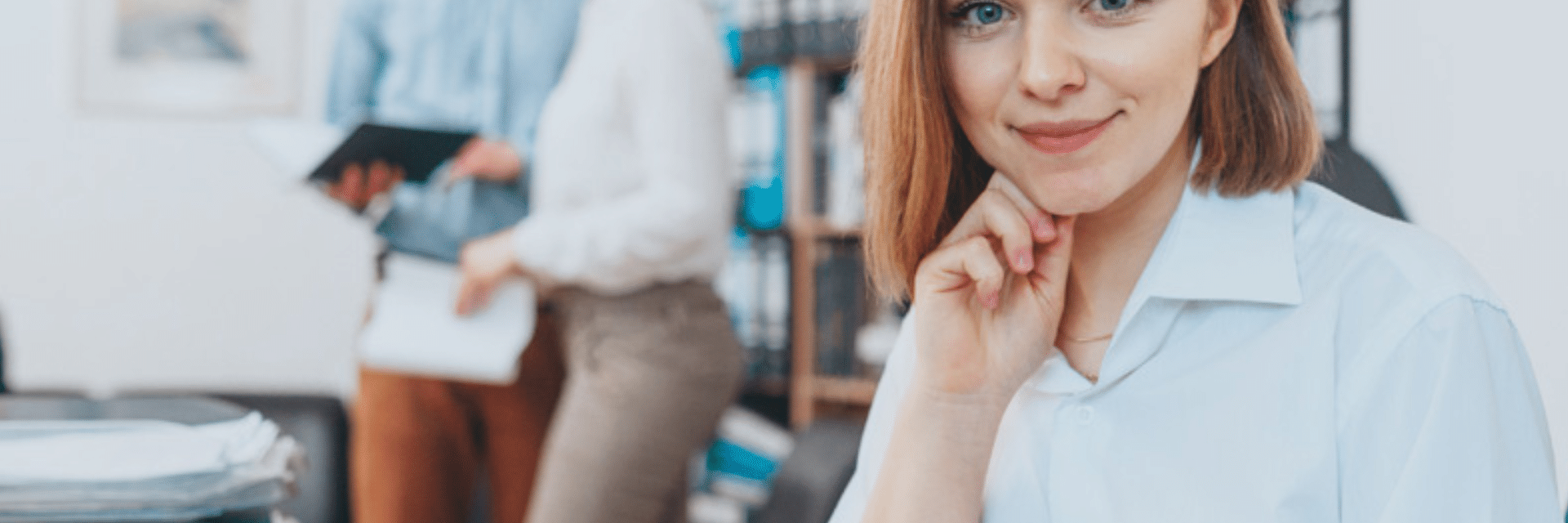 frau vor einem laptop. Im Hintergrund stehen ein Mann und eine Frau und sehen gemeinsam in ein buch.